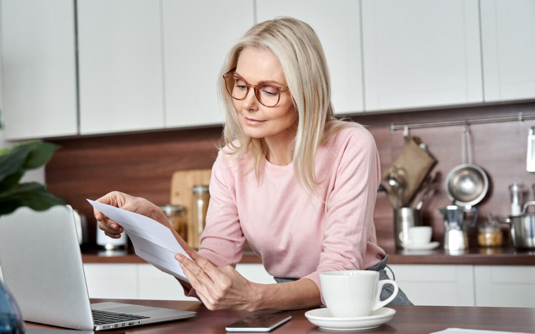 Una mujer con gafas lee un documento impreso en la mesa de su cocina, con un ordenador portátil abierto y una taza de café a su lado.