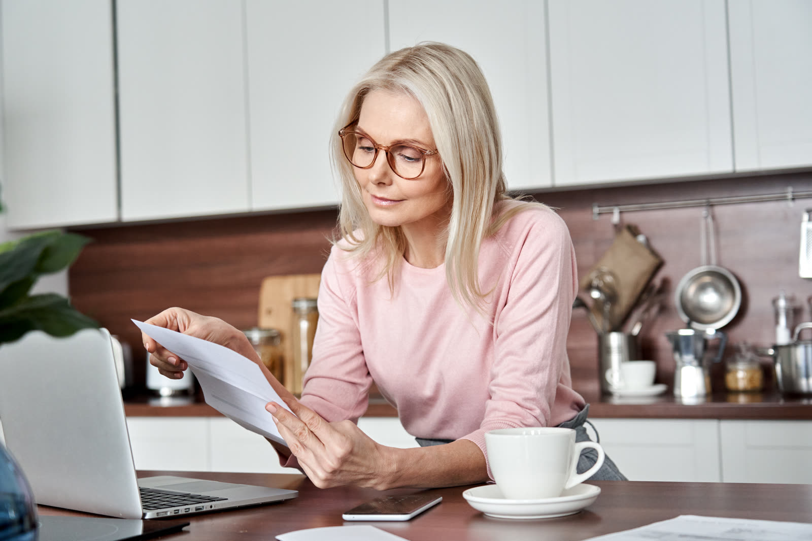 A woman in glasses reads a printed document at her kitchen table, with an open laptop and a cup of coffee beside her.